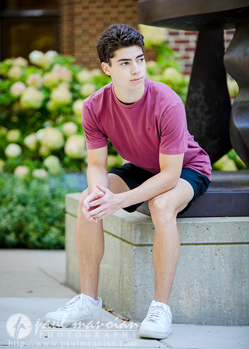 A young man with short dark hair, wearing a maroon T-shirt, dark shorts, and white sneakers, sits on a concrete bench outdoors during his Saline senior pictures session. Greenery and flowers are visible in the background.