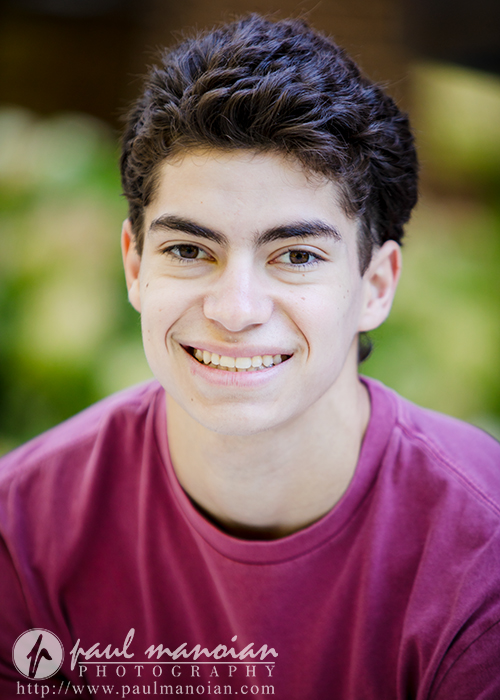 A young person with short, dark curly hair smiles at the camera during a Saline senior pictures session, wearing a maroon shirt. The softly blurred background reveals hints of greenery. A watermark for Paul Manoian Photography is visible at the bottom.