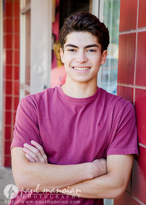 A teenage boy with short dark hair wearing a mauve t-shirt stands against a red-tiled wall, smiling with his arms crossed during a Saline senior pictures session in an outdoor urban setting.