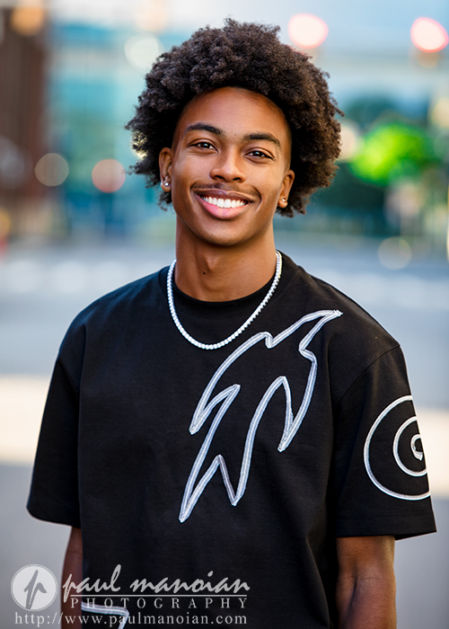 A young man with curly hair smiles at the camera while standing outside. Perfect for senior pictures Detroit, he wears a black shirt with white graphic designs and a silver chain necklace, the softly blurred background highlighting him as the focus.