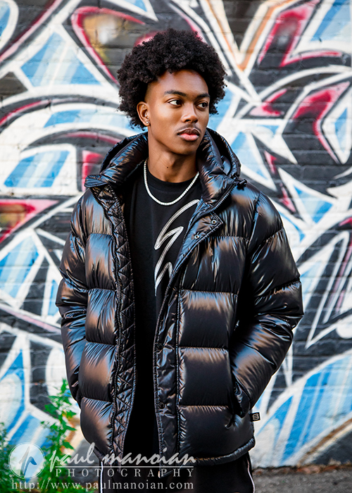 A young man wearing a shiny black puffer jacket stands in front of a colorful graffiti-covered brick wall, looking slightly to the side with his hands in his pockets—perfect for senior pictures Detroit style.