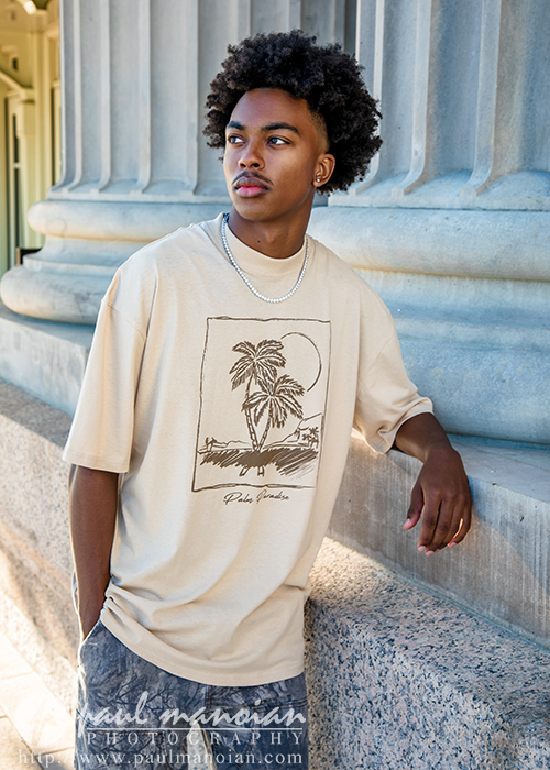 A young man with curly hair stands by a stone column in Detroit, wearing a beige T-shirt with a tropical palm tree graphic and gray shorts, looking off to the side with a thoughtful expression—perfect for senior pictures Detroit style.