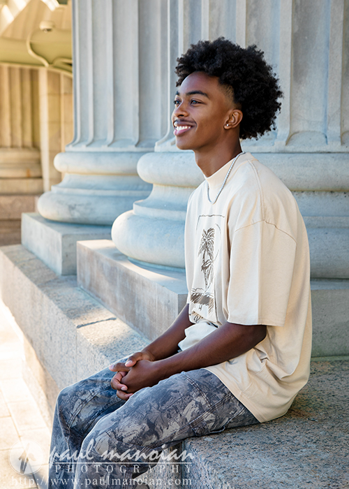 A young man with curly hair sits and smiles on stone steps in front of large columns, posing for his senior pictures in Detroit. He wears a light-colored T-shirt with a palm tree print and patterned pants.