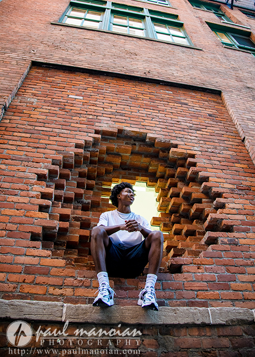 A young man sits in a circular hole in a brick wall, smiling and looking to the side during his senior pictures. He is wearing a white t-shirt, black shorts, and sneakers. Tall brick building windows are visible above him in Detroit.