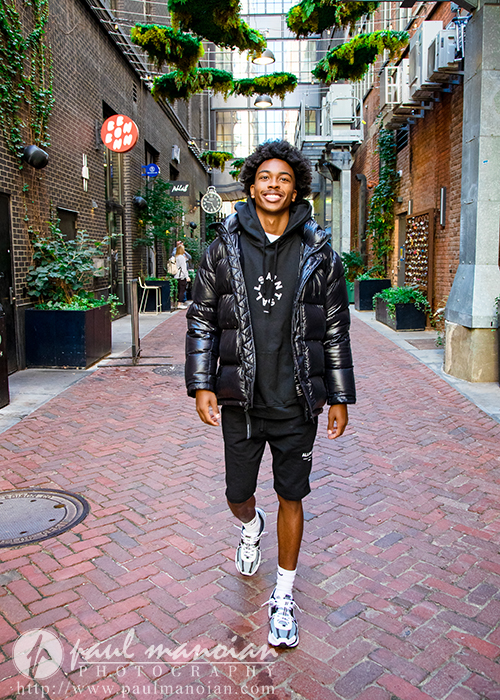 A young man wearing a black puffer jacket, black hoodie, black shorts, and white sneakers walks smiling down a brick alley lined with greenery and shops—perfect inspiration for senior pictures Detroit style.