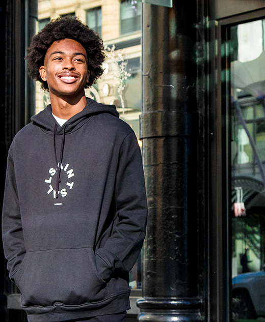 A young man with curly hair smiles while standing outside a building in Detroit, wearing a black hoodie with white text. Sunlight brightens the scene, perfect for senior pictures, and city reflections are visible in the windows behind him.