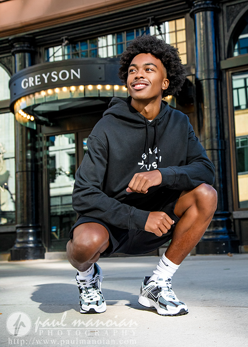A young man in a black hoodie and shorts squats and smiles outdoors in front of a building with a "GREYSON" sign overhead. Perfect for senior pictures Detroit style, he pairs white sneakers and socks as the sun casts shadows on the ground.