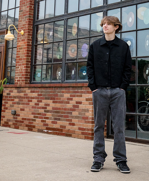 A young man in casual clothes stands on a sidewalk in front of a brick building adorned with colorful vinyl records—capturing a relaxed moment during his Milford senior pictures session. He looks to the side, hands tucked in his pockets.