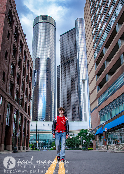 A young man in a red and black letterman jacket stands in the middle of an empty city street, tall modern skyscrapers and older brick buildings behind him during his St. Clair Shores senior pictures session.