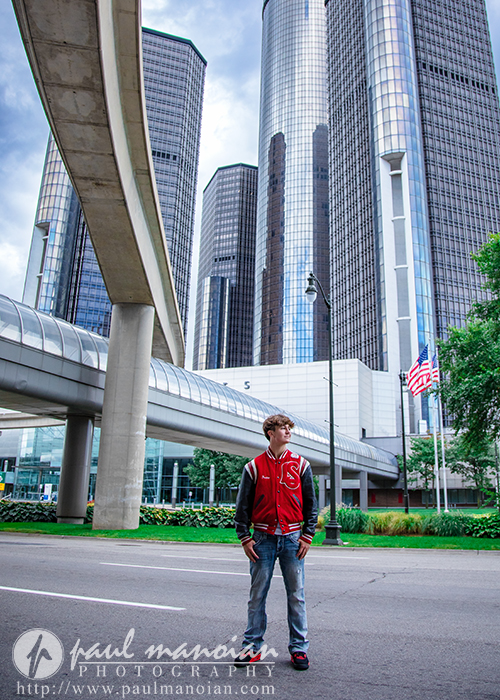 A young man in a red varsity jacket stands on a city street near a monorail track, with glass skyscrapers and an American flag in the background during his St. Clair Shores senior pictures session.