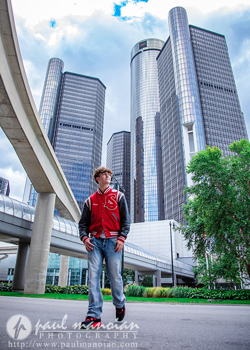 A young man in a red varsity jacket stands outdoors in front of modern glass skyscrapers, greenery, and elevated walkways under a partly cloudy sky during his St. Clair Shores senior pictures session.