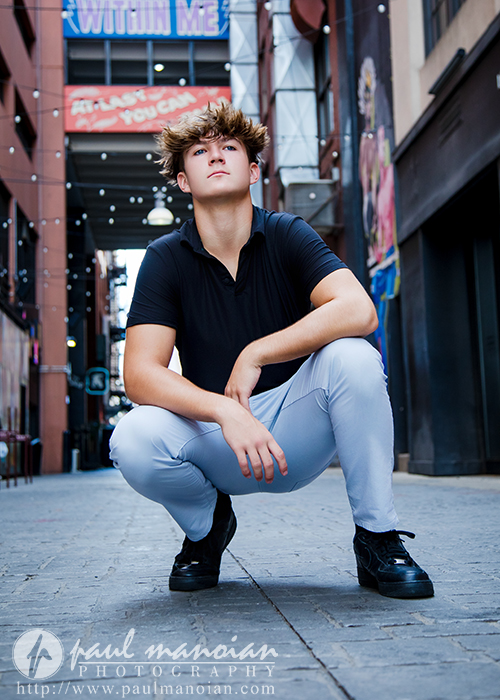 A young man with light brown hair crouches confidently in a city alleyway during his St. Clair Shores senior pictures session, wearing a black polo shirt, light gray pants, and black shoes. Bright signs and string lights decorate the urban background.