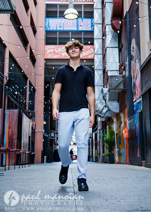 A young man wearing a black shirt and white pants walks confidently down a colorful urban alleyway, murals and string lights illuminating his St. Clair Shores senior pictures session, with a large "WITHIN ME" sign above him.