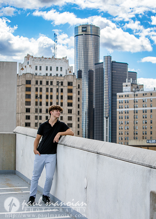A young man with curly hair, wearing a black shirt and white pants, leans on a rooftop ledge with tall city buildings and a partly cloudy sky in the background during his St. Clair Shores senior pictures session.