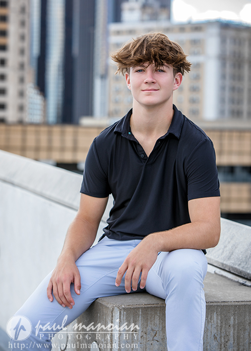 A teenage boy with light brown hair, wearing a black polo shirt and light blue pants, sits on a concrete ledge during his St. Clair Shores senior pictures session, with a city skyline in the background.