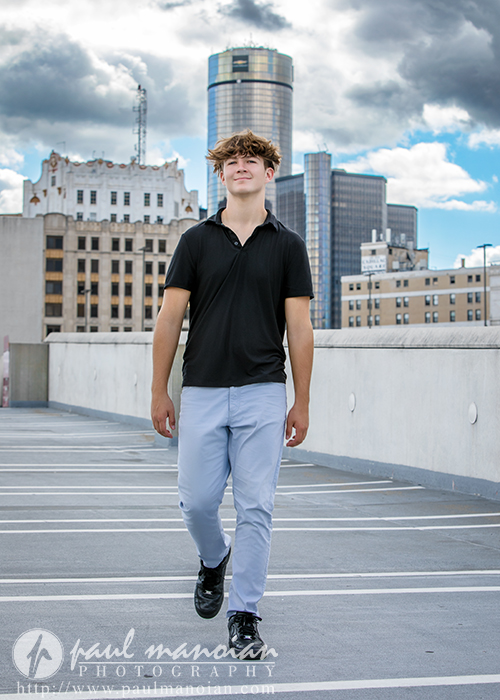 A young man with brown hair, wearing a black shirt and light blue pants, walks confidently in a rooftop parking lot during his St. Clair Shores senior pictures session, with city buildings and a cloudy sky in the background.