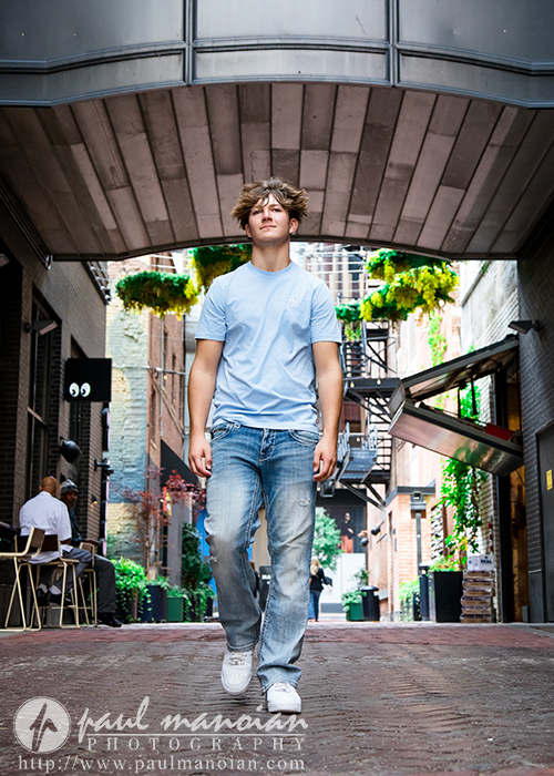 A young man in a light blue t-shirt and jeans walks confidently down a brick alleyway lined with plants and outdoor seating, with modern architecture overhead—a perfect setting for a St. Clair Shores senior pictures session.
