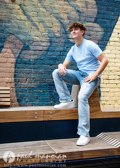 A young man in a light blue t-shirt and jeans poses confidently on a wooden bench in front of a colorful brick wall mural during his St. Clair Shores senior pictures session.
