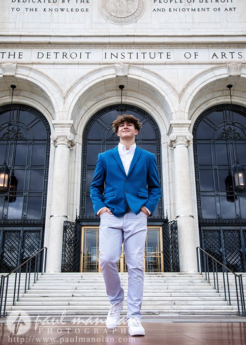 A young man in a blue blazer and white pants stands confidently on steps in front of the Detroit Institute of Arts, showcasing his style during a St. Clair Shores senior pictures session, with grand arched doorways and columns behind him.