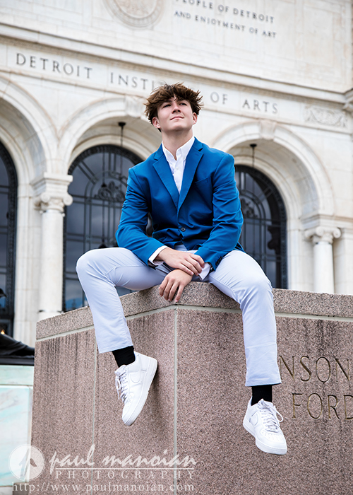 A young man in a blue blazer and white pants sits confidently on a stone ledge outside the Detroit Institute of Arts, capturing a stylish moment during his St. Clair Shores senior pictures session, with arched windows and engraved text in the background.