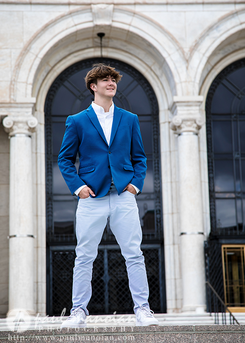 A young man in a bright blue blazer, white shirt, and light pants stands confidently outdoors in front of a grand building with arched doorways and columns during his St. Clair Shores senior pictures session, hands in his pockets and smiling slightly.
