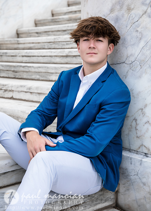 A young man with light brown hair, wearing a blue blazer and white pants, sits on marble steps during his St. Clair Shores senior pictures session, leaning against a marble wall and looking confidently at the camera.