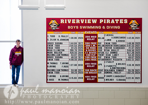 A high school senior in a Riverview Pirates hoodie stands by large windows beside the boys swimming and diving record board, captured during his Riverview senior pictures session, highlighting event records, swimmers, and years.