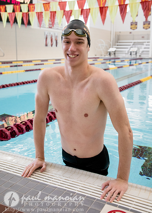 A young male swimmer in a swim cap and goggles smiles while standing at the edge of an indoor pool during his Riverview senior pictures session, with colorful lane dividers and banners in the background.