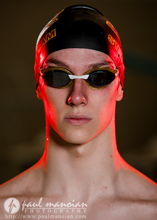 A swimmer wearing a swim cap and goggles stares directly at the camera, illuminated by dramatic red lighting during a Riverview senior pictures session. The image is watermarked with "paul manoian photography.