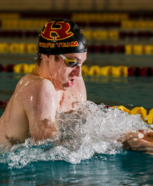 A swimmer wearing goggles and a swim cap labeled "SWIM TEAM" competes in the pool, performing the breaststroke amid splashing water—an energetic scene perfect for Swimming Senior Pictures, with yellow and red lane dividers behind.