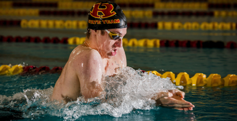 A swimmer wearing goggles and a swim cap labeled "SWIM TEAM" competes in the pool, performing the breaststroke amid splashing water—an energetic scene perfect for Swimming Senior Pictures, with yellow and red lane dividers behind.