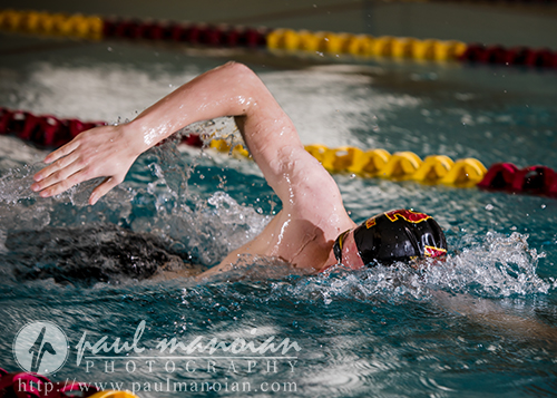 A swimmer, possibly prepping for their Riverview senior pictures session, wears a black cap and goggles while performing a freestyle stroke in an indoor pool with red and yellow lane markers. Water splashes around their arm as they move forward.