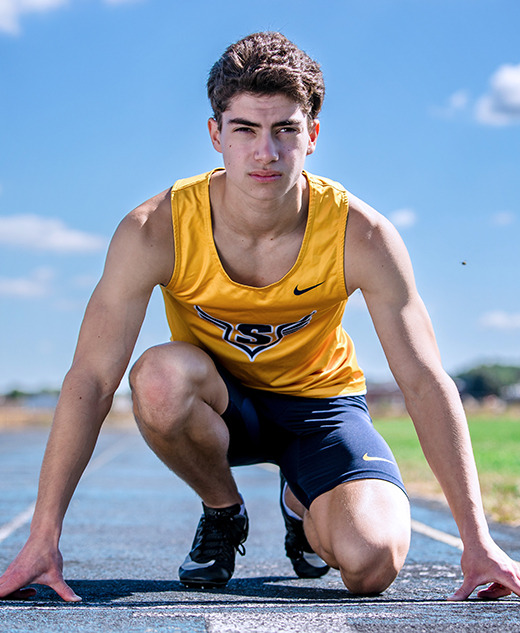 A young male athlete in a yellow tank top and navy shorts crouches at the starting line of an outdoor track, poised to sprint under a clear blue sky—an action moment from his Saline senior pictures session.