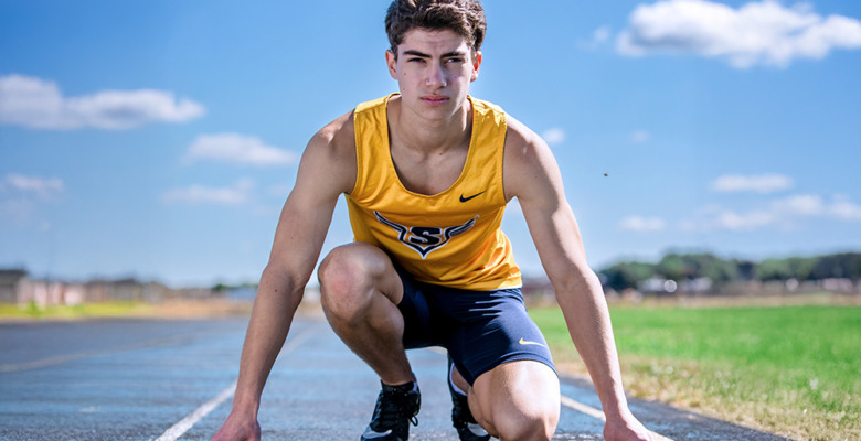 A young male athlete in a yellow tank top and navy shorts crouches at the starting line of an outdoor track, poised to sprint under a clear blue sky—an action moment from his Saline senior pictures session.