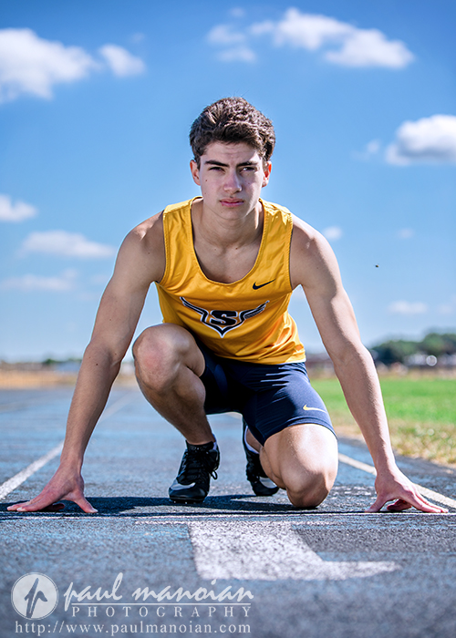 A male athlete in a yellow and navy uniform is crouched in a starting position on an outdoor track during his track senior pictures session, looking focused under a bright blue sky with scattered clouds.