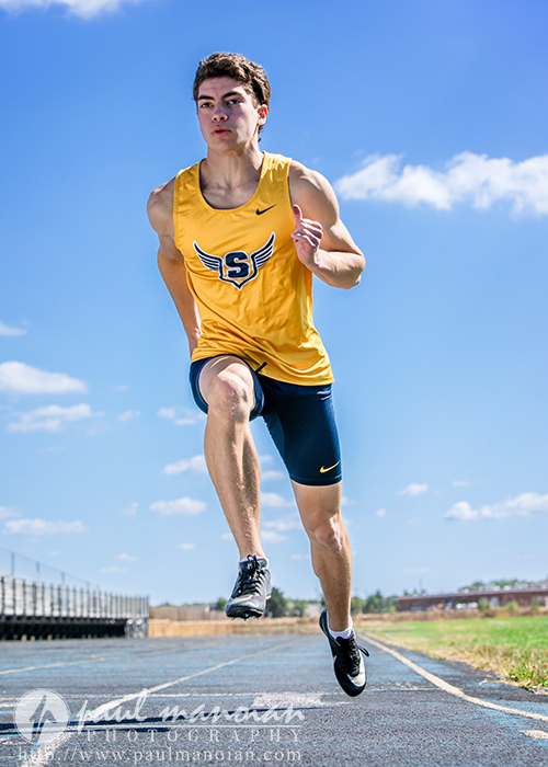 A male athlete wearing a yellow tank top with a winged "S" logo and navy shorts runs on an outdoor track under a blue sky. Captured mid-stride during his track senior pictures session, he looks focused and determined.