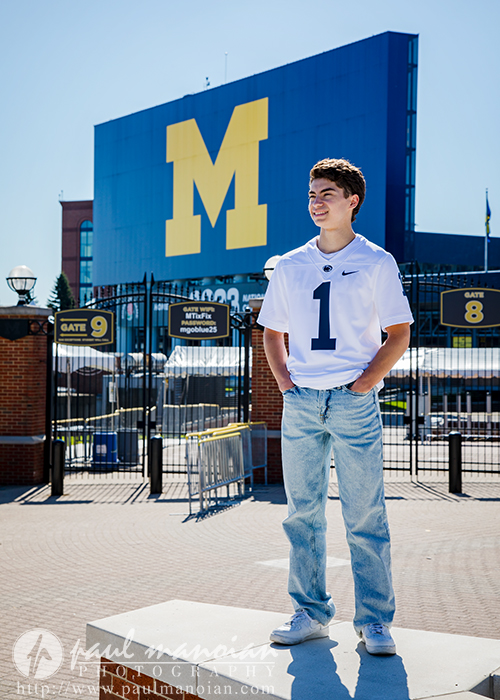 A young man in a white jersey and jeans stands smiling in front of a large blue and yellow "M" sign, capturing his senior pictures at Michigan Stadium's entrance.