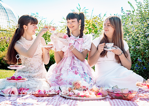 Three high school senior girls in pastel dresses enjoy tea and sweets at an outdoor vintage tea party. The scene is bright and cheerful, perfect for a senior pictures session with desserts, teacups, and flowers set on a blanket in a garden setting.