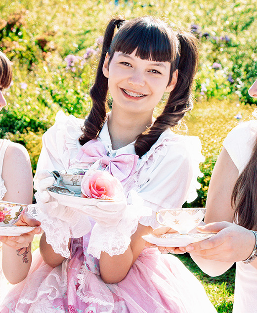 Three young high school seniors in light-colored, vintage-inspired dresses are smiling and holding decorative teacups at a sunny garden vintage tea party senior pictures session. One holds a plate with a pink rose as they enjoy the afternoon together.