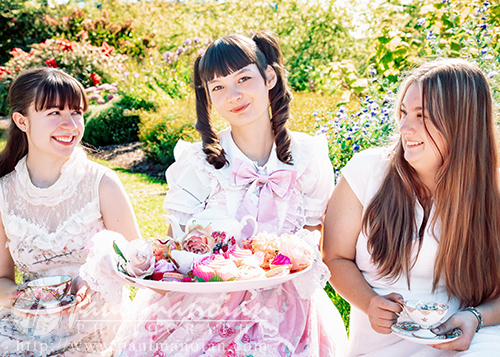 Three high school senior girls sit outdoors, smiling and holding teacups during a vintage tea party senior pictures session. The woman in the center wears a pink and white frilly dress and holds a tray of colorful cupcakes amid bright flowers and greenery.