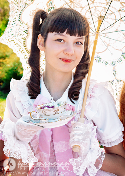 A high school senior girl in a pink and white dress holds a decorative teacup and saucer, posing for vintage tea party senior pictures. With curled pigtails, lace gloves, and a smile, she stands under a lace parasol outdoors.