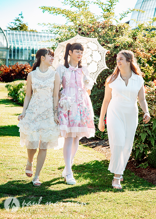 Three high school senior girls in light, elegant dresses walk together in a sunny garden during a vintage tea party senior pictures session. One holds a white lace parasol, and they smile as they enjoy each other's company amid the greenery and flowers.