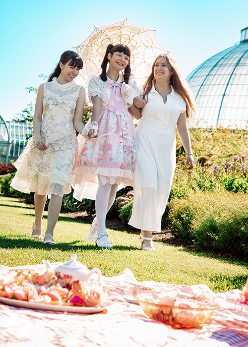 Three high school senior girls in light, elegant dresses walk together in a sunny garden, smiling. One holds a lacy parasol. In the foreground, a vintage tea party picnic with tea and pastries is laid out—perfect for a senior pictures session. Glass domes appear behind them.