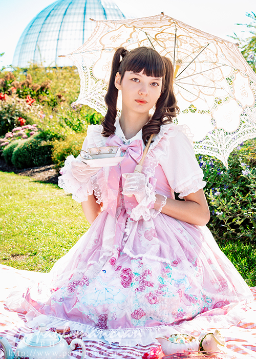 A high school senior girl in a frilly pink dress sits on a picnic blanket in a garden, holding a lace parasol and teacup—a perfect vintage tea party scene for senior pictures. She is surrounded by lush greenery, flowers, and a glass dome in the background.