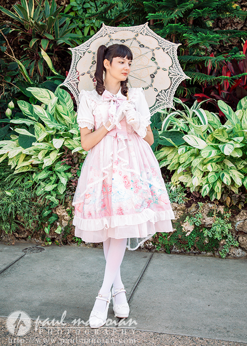 A high school senior girl in a pink and white frilly dress holds a lace parasol, styled for a vintage tea party senior pictures session. She stands on a path with lush green foliage, wearing white tights and shoes in classic fashion.