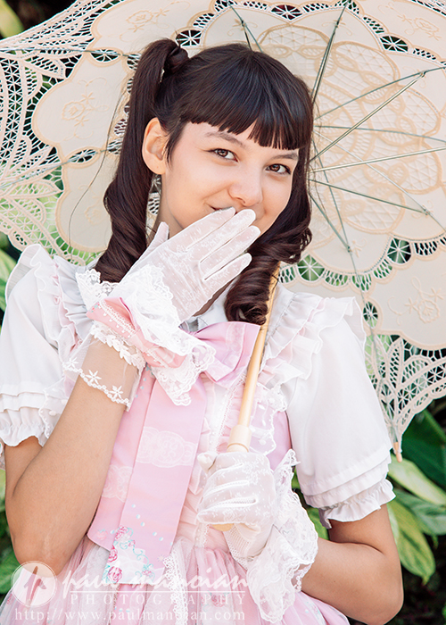 A high school senior girl in a pink and white frilly dress holds a lace parasol and covers her mouth with a gloved hand, smiling playfully. She stands outdoors, surrounded by greenery—perfect for a vintage tea party senior pictures session.