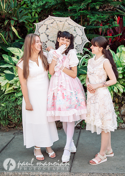 Three high school senior girls in white and pastel dresses stand together outdoors, smiling and laughing during a vintage tea party senior pictures session. The woman in the center holds a lacy parasol, with lush green plants filling the background.
