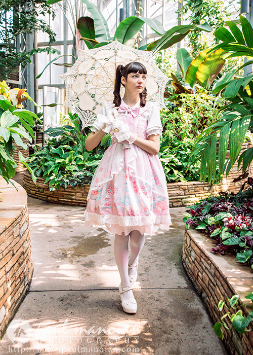 A high school senior girl in a pastel pink dress holds a lace parasol while standing on a garden path, surrounded by lush greenery in a greenhouse—perfect for a vintage tea party or whimsical senior pictures session.
