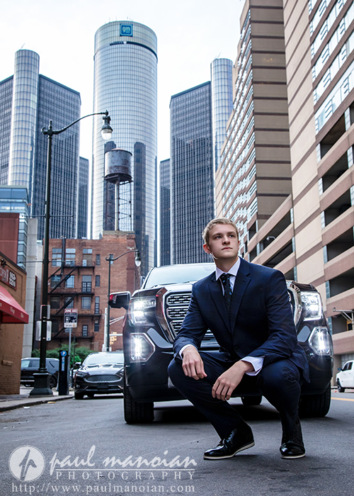 A young man in a suit kneels on one knee in front of a black SUV on a city street during a session for White Lake senior pictures in Detroit, with tall modern buildings and the GM Renaissance Center rising in the background.