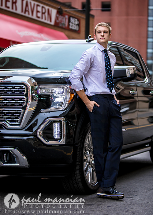 A young man in a dress shirt, tie, and slacks leans casually against a shiny black pickup truck on a city street during his session for White Lake senior pictures in Detroit, with a restaurant sign visible in the background.
